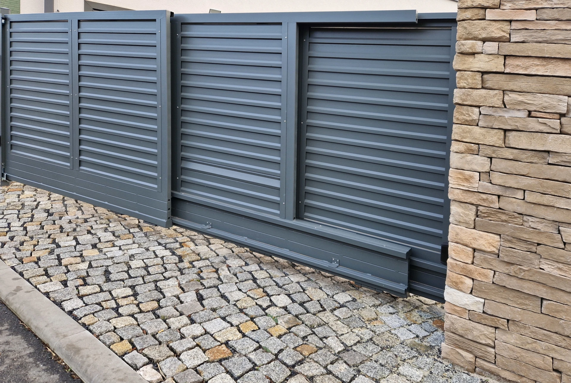 Entrée résidentielle avec portail coulissant automatique en aluminium anthracite à lames horizontales, intégré dans clôture maçonnée blanc cassé avec piliers béton