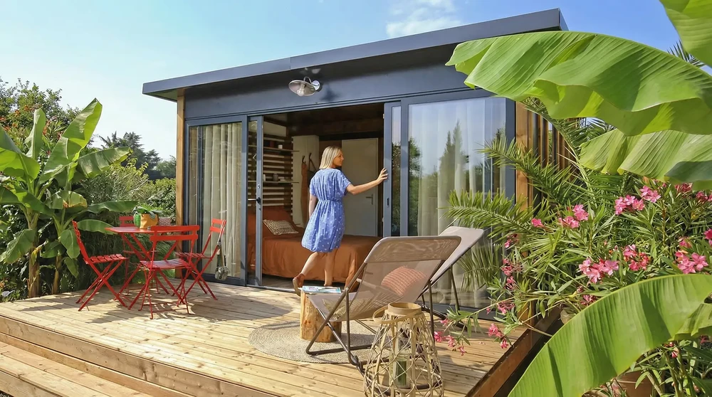 Femme blonde en robe bleue entrant dans un studio de jardin moderne, terrasse en bois composite, mobilier de jardin rouge (table et chaises bistrot), fauteuil de relaxation textilène